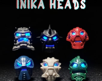 Può includere: Sei teste Inika della serie Voya-Nui, ognuna con occhi blu luminosi. Le teste sono di vari colori: grigio, nero, rosso, blu, bianco e verde. Il testo "INIKA HEADS" è visualizzato sopra le teste.