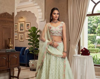 Op de afbeelding: Een lichtgroene geborduurde lehenga met bloemendetails. De outfit bevat een bijpassende top en een doorschijnende dupatta. De lehenga heeft roze bloemenborduurwerk langs de zoom en een bijpassend bloemmotief op de top.
