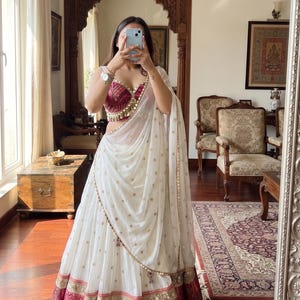 May include: A white and magenta lehenga set featuring a sleeveless blouse with intricate patterns and a pleated skirt with a wide magenta border. A sheer white dupatta with gold circular accents and a gold-trimmed edge drapes over the shoulder.