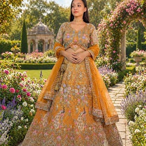 May include: Mustard yellow embroidered lehenga with peacock and floral motifs. The outfit includes a matching blouse and dupatta. The intricate embroidery features pink, blue, and gold accents. The lehenga is worn outdoors in a garden setting.