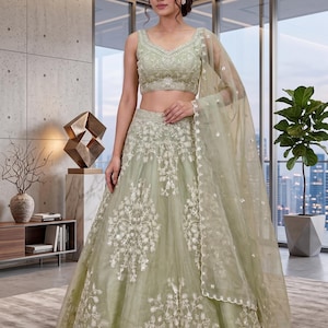 May include: A sage green lehenga choli set featuring a sleeveless blouse and a floor-length skirt, both adorned with intricate white floral embroidery and delicate sequin detailing. A matching sheer dupatta with a scalloped border is draped over one shoulder.