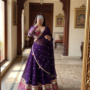 May include: A deep purple lehenga choli set featuring a long, flowing skirt with gold polka dot embroidery and a wide, multicolored patterned border at the hem. The matching long-sleeved blouse has intricate embroidery and a sweetheart neckline.