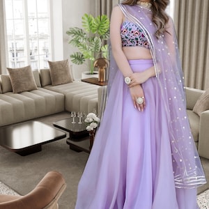 Pode incluir: Uma mulher vestindo um traje formal lavanda. O traje inclui um top bordado floral, uma saia longa e um lenço transparente com detalhes dourados. A palavra "Fashion" está no canto superior esquerdo.