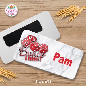 May include: A rectangular magnet with a white marble design, featuring red dice and the text "It's Bunco Time!" and "Pam". A second magnet is visible behind it. The style is N65.