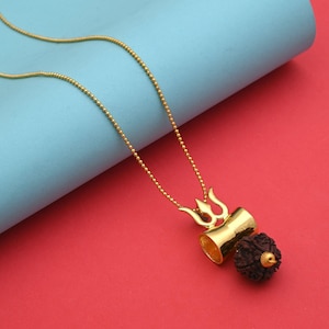May include: A gold-coloured pendant necklace. The pendant features a trident design, a cylindrical element, and a dark brown, textured bead. The necklace hangs on a delicate chain against a red and blue background. The pendant is likely made of metal.