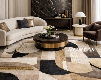 Op de afbeelding: Een moderne woonkamer met een crèmekleurige bank, een donkerbruine salontafel met een gouden band en een patroonkleed. Het kleed heeft een geometrisch ontwerp in beige, zwart en goud. De tekst "Ambar rugs" is zichtbaar op het kleed.
