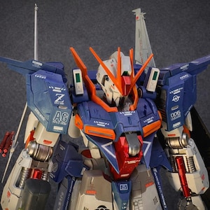May include: A detailed model of a mecha robot, featuring a color scheme of blue, orange, white, and red. The robot has intricate paneling, wings, and various mechanical components. The model is a collectible item.