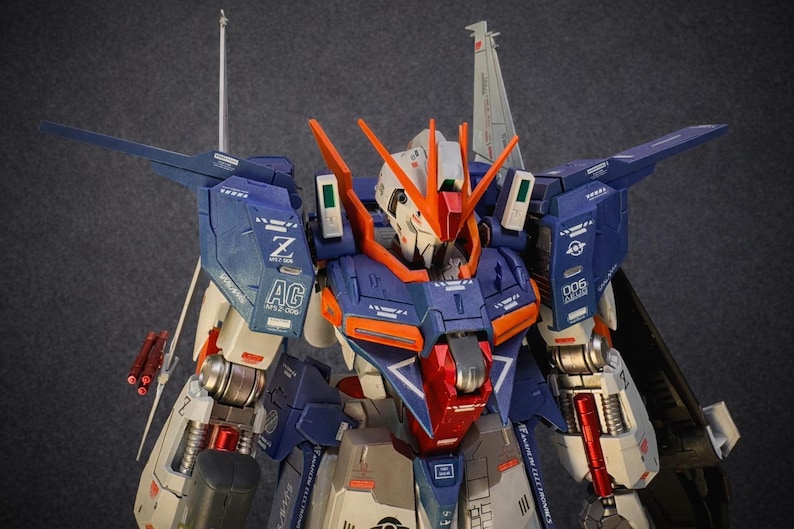 May include: A detailed model of a mecha robot, featuring a color scheme of blue, orange, white, and red. The robot has intricate paneling, wings, and various mechanical components. The model is a collectible item.