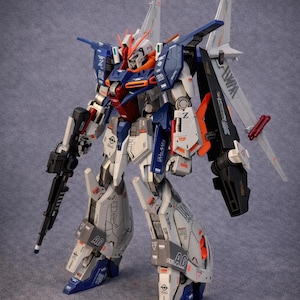 May include: A detailed model of a robot with a white, blue, orange, and gray color scheme. The robot has a gun in its left hand and large wings on its back. The model is highly detailed with intricate panel lines and markings.