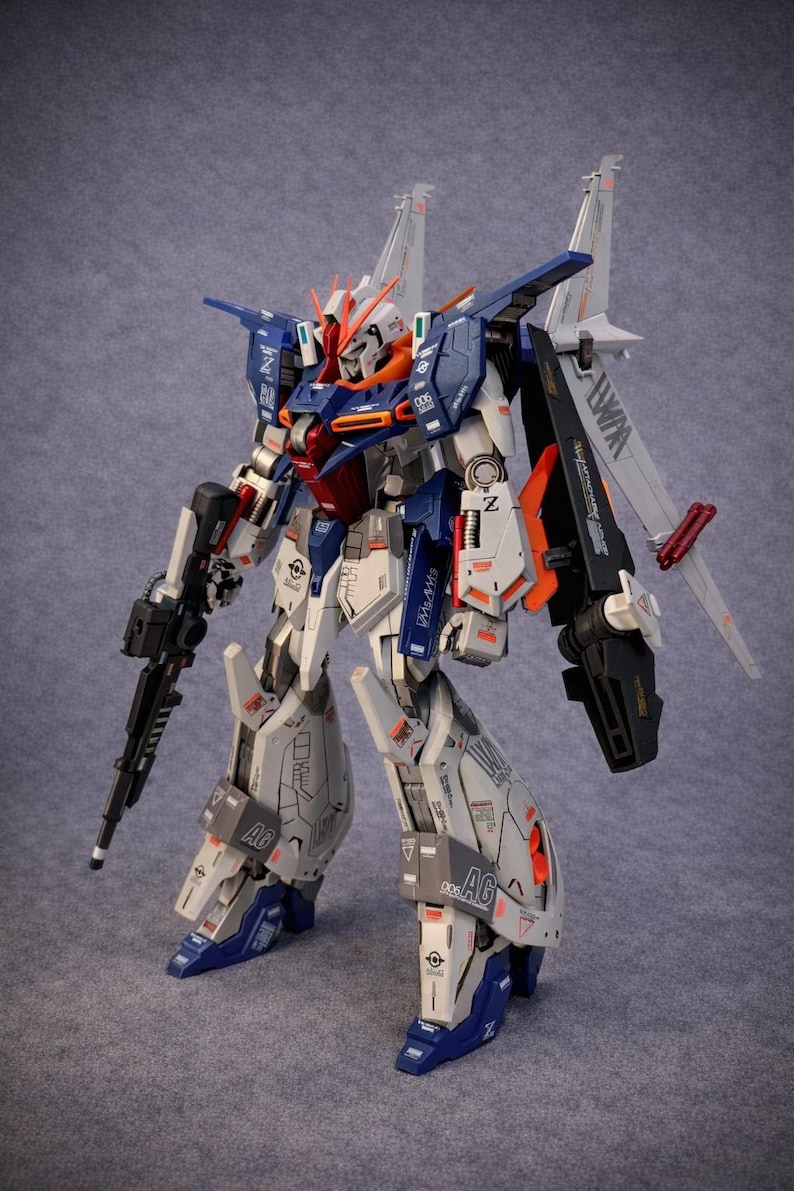 May include: A detailed model of a robot with a white, blue, orange, and gray color scheme. The robot has a gun in its left hand and large wings on its back. The model is highly detailed with intricate panel lines and markings.