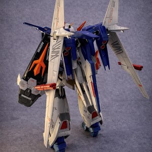 May include: A detailed model of a robot with white, blue, and orange accents. The robot has large wings and is standing on a gray surface. The model has the text "WIM" and "AGC1006" printed on it.