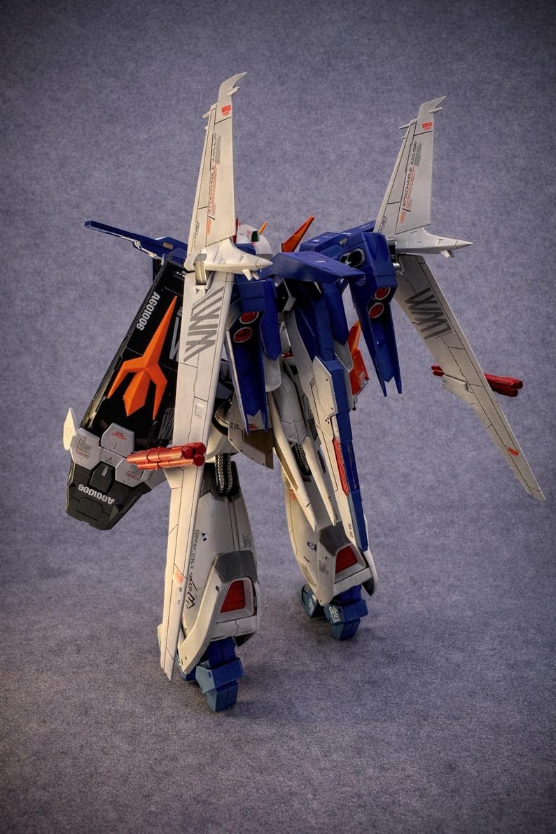 May include: A detailed model of a robot with white, blue, and orange accents. The robot has large wings and is standing on a gray surface. The model has the text "WIM" and "AGC1006" printed on it.