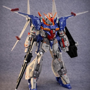May include: A detailed model of a mecha robot, featuring a color scheme of blue, white, gray, orange, and red. The robot has multiple articulated parts, wings, and a large rifle. The model is standing upright against a gray background.