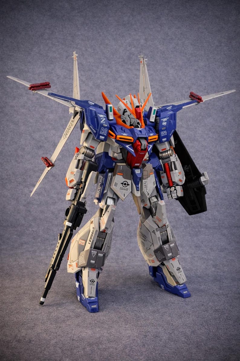 May include: A detailed model of a mecha robot, featuring a color scheme of blue, white, gray, orange, and red. The robot has multiple articulated parts, wings, and a large rifle. The model is standing upright against a gray background.