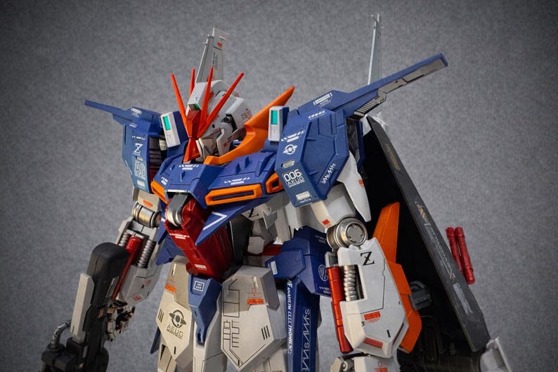 May include: A detailed model of a robot with a blue, white, orange, and red color scheme. The robot has intricate mechanical details, including panels, joints, and weapons. The model is posed against a gray background.