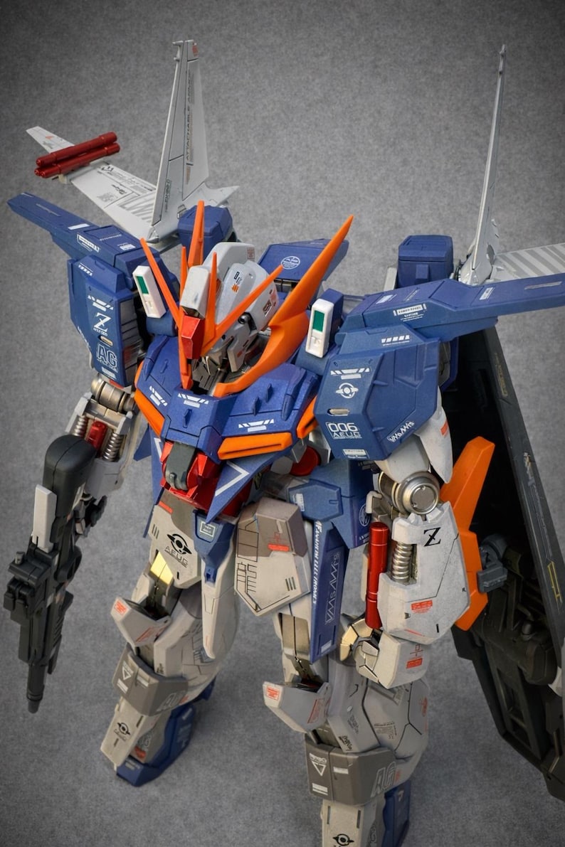 May include: A detailed model of a mecha robot, featuring a color scheme of blue, orange, gray, and white. The robot has intricate mechanical details, including wings, weapons, and various panels. The model is a collectible item.
