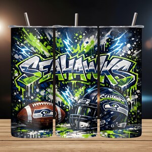 May include: A stainless steel tumbler featuring a Seattle Seahawks design. The design includes the team's logo, a football, and a helmet, all set against a backdrop of blue, green, and white graffiti-style artwork. The tumbler has a silver straw.