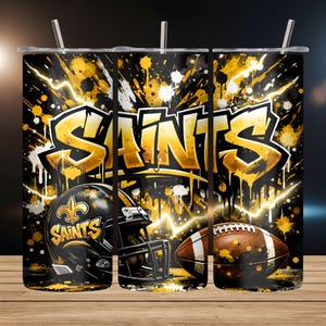 May include: A stainless steel tumbler featuring a bold design with the word "SAINTS" in gold graffiti style. The design includes a football helmet, a football, and gold and white paint splatters on a black background. The tumbler has a silver straw.
