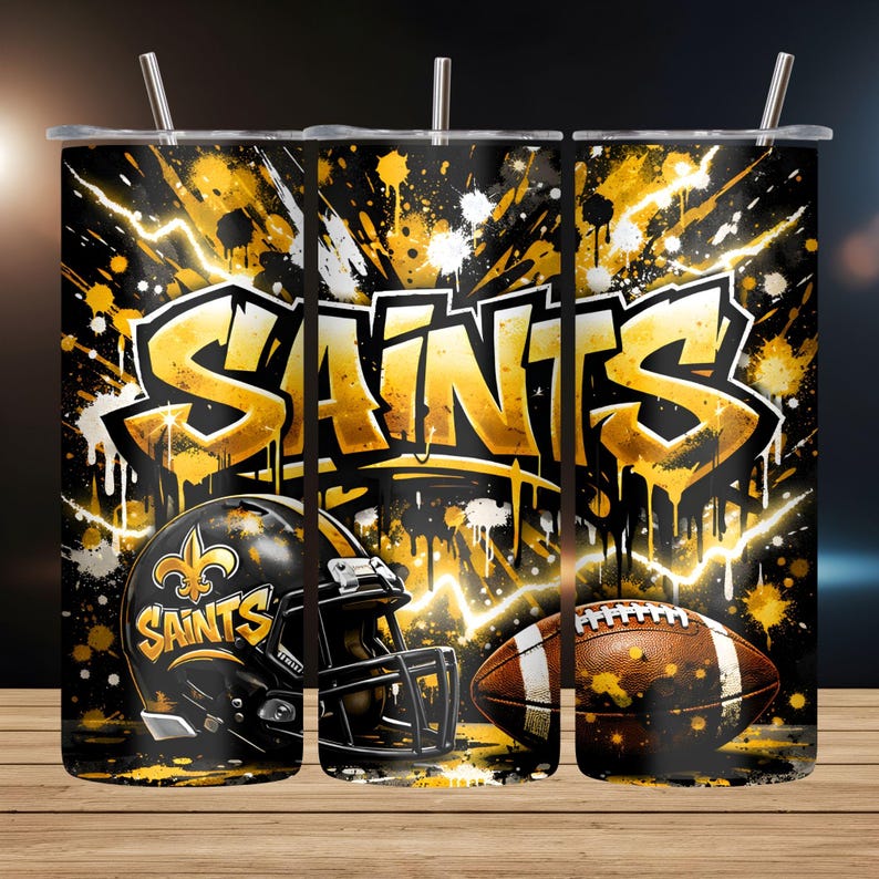 May include: A stainless steel tumbler featuring a bold design with the word "SAINTS" in gold graffiti style. The design includes a football helmet, a football, and gold and white paint splatters on a black background. The tumbler has a silver straw.