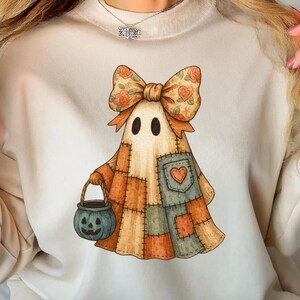 Op de afbeelding: Crèmekleurige sweatshirt met een grillig spookontwerp. Het spook heeft een patchworkpatroon in oranje, bruine en blauwe tinten, een bloemenstrik en een teal pompoenemmer. Het ontwerp heeft een charmante, handgetekende esthetiek, perfect voor Halloween.