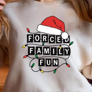 May include: Cream-colored sweatshirt with a graphic that reads "FORCED FAMILY FUN" in black block letters. A red Santa hat with white trim sits above the text. The text is surrounded by a string of colorful Christmas lights.