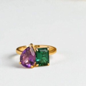 Op de afbeelding: Een gouden ring met een peervormige paarse edelsteen en een smaragdgroene vierkante edelsteen. De ring staat op een witte achtergrond, waardoor de levendige kleuren van de edelstenen opvallen.