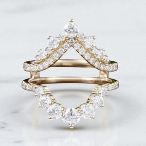 May include: A gold-colored ring guard with a V-shaped design, encrusted with numerous small, clear gemstones. The ring guard has a curved bottom row of larger gemstones and a delicate, detailed setting.