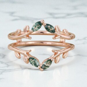 Puede incluir: Anillo de oro rosa con un diseño de doble banda. El anillo presenta detalles en forma de hojas y piedras preciosas verdes. Las piedras preciosas están engastadas en el centro del anillo y los detalles de hojas están en los lados.
