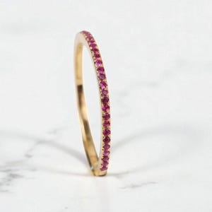 Op de afbeelding: Een gouden ring met kleine, helderrode edelstenen. De ring is dun en delicaat, met de edelstenen dicht op elkaar gezet rond de band. De ring wordt getoond op een wit marmeren oppervlak.