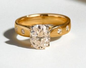May include: A gold ring with a large oval-cut diamond center stone. The band features small, round accent stones. The ring is set on a white surface, with shadows adding depth. A classic piece of jewelry.