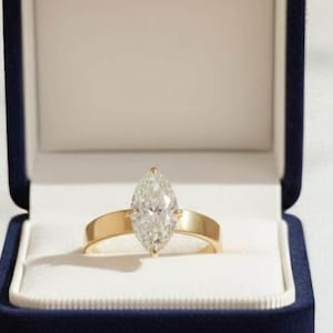 May include: A marquise-cut diamond ring with a gold band, presented in a navy blue velvet ring box with a white interior. The ring is centered in the box, showcasing its elegant design.