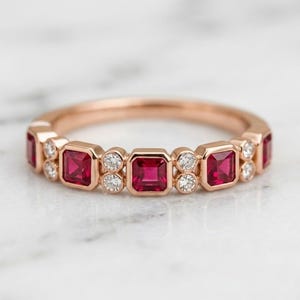 May include: A rose gold band ring featuring a series of alternating square-cut red rubies and small round white diamonds. The gemstones are set in a bezel style along the top half of the ring, creating a continuous pattern of red and clear stones.