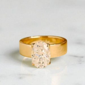 Op de afbeelding: Een gouden ring met een grote, ovaalvormige, lichtgeleurde edelsteen. De ring heeft een brede, platte band. De edelsteen is gezet in een eenvoudige vier-klauwzetting. De ring wordt getoond op een wit oppervlak.