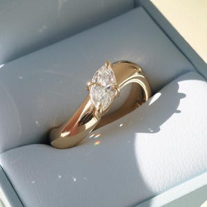 Könnte beinhalten: Ein goldener Ring mit einem Diamanten im Marquiseschliff, der in einem polierten Band gefasst ist. Der Ring wird in einer hellblauen Schmuckschatulle präsentiert, wobei sich das Sonnenlicht auf der Oberfläche des Rings spiegelt. Das Design des Rings ist modern und elegant.