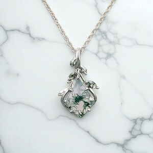 Op de afbeelding: Een zilveren hangerketting met een edelsteen met groene en witte insluitsels. De hanger heeft een decoratief zilveren frame met bladvormige accenten en hangt aan een fijne zilveren ketting. De ketting wordt getoond op een wit marmeren oppervlak.