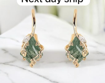 May include: Gold-coloured earrings featuring a green gemstone centre. The earrings have a leaf design and a small, clear stone. The text "Next day ship" is in the background.