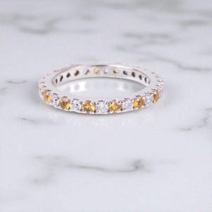 Könnte beinhalten: Ein silberner Eternity-Ring mit abwechselnd gelben und weißen Edelsteinen. Der Ring liegt auf einer weißen Marmoroberfläche, die das Funkeln der Edelsteine zur Geltung bringt. Ein filigranes Schmuckstück.
