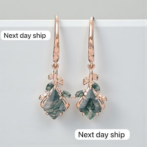 May include: A pair of rose gold earrings with a leaf design and green gemstone pendants. The earrings have a long, slender hook and a textured leaf detail. The gemstones are a light green color with darker green inclusions.