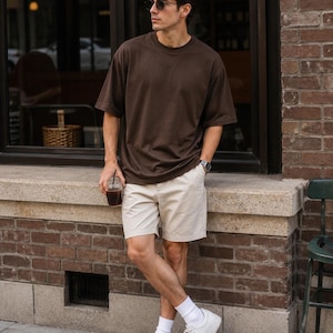 May include: A dark brown short-sleeved crew neck t-shirt, beige chino shorts, white crew socks, and white low-top sneakers. The outfit is completed with dark-tinted sunglasses and a silver-tone wristwatch worn on the left wrist.