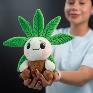 May include: A plush toy featuring a smiling, cartoon-like character with a white face, green leaf-like hair, and a brown body. The toy has small arms and legs, and pink blush marks on its cheeks. The toy is being held in front of a dark background.