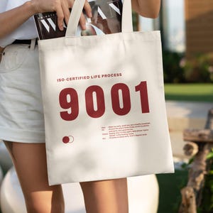 Puede incluir: Bolso tote color crema con el texto "ISO-CERTIFIED LIFE PROCESS" y el número "9001" en rojo. La bolsa también presenta una lista de instrucciones y un pequeño gráfico. La bolsa es sostenida por una persona.