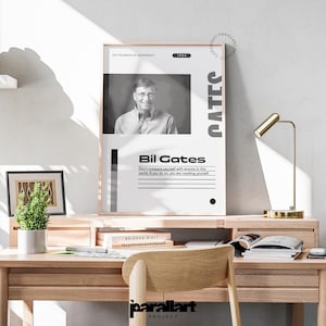 May include: A framed poster featuring a black and white image of Bill Gates, co-founder of Microsoft, with the text "Bil Gates" and a quote. The poster is displayed on a wooden desk with a small plant and a gold desk lamp.