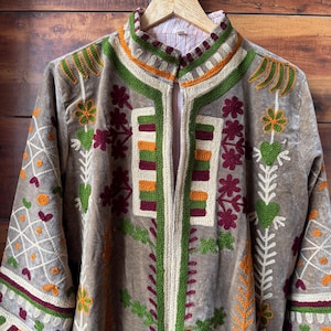 Puede incluir: Chaqueta de terciopelo marr&oacute;n con intrincados bordados en verde, naranja y burdeos. La chaqueta presenta un cuello mandar&iacute;n y un dise&ntilde;o vertical en la parte delantera. Las mangas largas tambi&eacute;n est&aacute;n bordadas. La chaqueta est&aacute; colgada en una percha de madera.