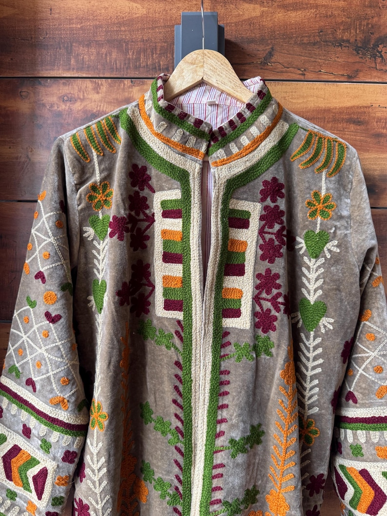 Puede incluir: Chaqueta de terciopelo marr&oacute;n con intrincados bordados en verde, naranja y burdeos. La chaqueta presenta un cuello mandar&iacute;n y un dise&ntilde;o vertical en la parte delantera. Las mangas largas tambi&eacute;n est&aacute;n bordadas. La chaqueta est&aacute; colgada en una percha de madera.