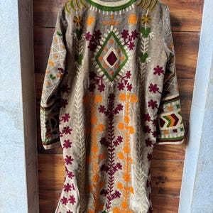 Puede incluir: Chaqueta larga bordada marr&oacute;n con dise&ntilde;o floral. La chaqueta presenta bordados en naranja, verde y granate. La chaqueta est&aacute; colgada en una percha de madera sobre un fondo de madera.