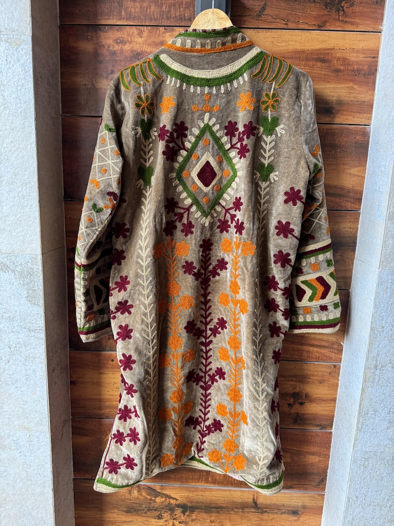 Puede incluir: Chaqueta larga bordada marr&oacute;n con dise&ntilde;o floral. La chaqueta presenta bordados en naranja, verde y granate. La chaqueta est&aacute; colgada en una percha de madera sobre un fondo de madera.