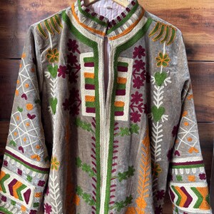 Puede incluir: Una chaqueta larga de color topo con intrincados bordados en verde, naranja y burdeos. La chaqueta presenta un cuello alto y una cremallera vertical. Patrones florales y geom&eacute;tricos adornan las mangas y el frente.