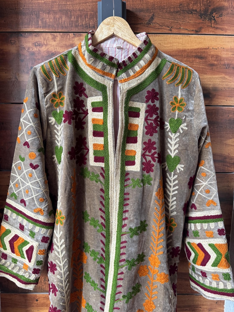 Puede incluir: Una chaqueta larga de color topo con intrincados bordados en verde, naranja y burdeos. La chaqueta presenta un cuello alto y una cremallera vertical. Patrones florales y geom&eacute;tricos adornan las mangas y el frente.