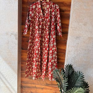 May include: A long, red dress with a white floral pattern hangs on a wooden hanger. The dress has a V-neck and three-quarter sleeves. The background is a wooden wall.