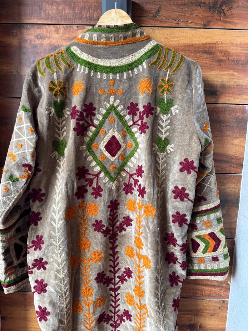 Puede incluir: Chaqueta de terciopelo color beige con intrincados bordados en tonos verdes, naranjas y burdeos. El dise&ntilde;o presenta patrones florales y geom&eacute;tricos, con un motivo en forma de diamante en la espalda. La chaqueta tiene mangas largas y cuello.